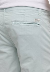 Light blue trousers with a back welt pocket and a small fabric label reading "INDICODE Carry Our Passion" near the waistband.