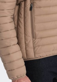 Beige pufferjakke med vandret quiltet mønster, sidelomme med lynlås og elastiske manchetter. Textureret stof med en glat overflade.