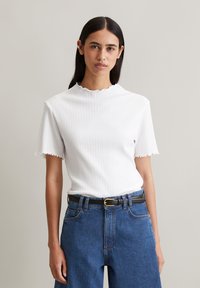 Weißes, geripptes Kurzarmoberteil mit Rüschenausschnitt, kombiniert mit hochgeschnittenen blauen Jeans und einem schwarzen Gürtel.