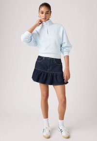 Γυναίκα που φοράει ανοιχτό μπλε πουλόβερ με φερμουάρ μέχρι τη μέση, σκούρο μίνι φούστα από τζιν με βολάν, άσπρες κάλτσες και άσπρα αθλητικά παπούτσια με μπλε και κόκκινες ρίγες.