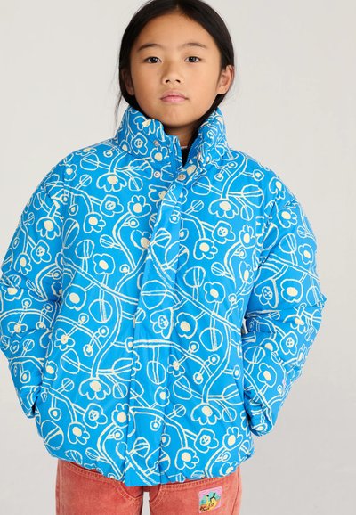 Enfant portant une doudoune bleue avec des lignes florales blanches et un pantalon rose, debout les mains dans les poches de la doudoune sur un fond uni.