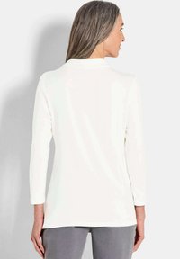 Maglia a maniche lunghe in morbido tessuto bianco, caratterizzata da un design semplice con colletto e orlo dritto, abbinata a pantaloni grigi.