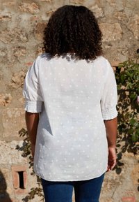 Persona con capelli ricci che indossa una maglietta bianca a maniche corte a pois e jeans blu, in piedi contro un muro di pietra con vegetazione.