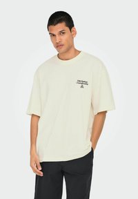 Camiseta extragrande de color crema hecha de algodón, con un estampado gráfico negro que dice "Campeonato de Palm Springs" en el área del bolsillo del pecho.