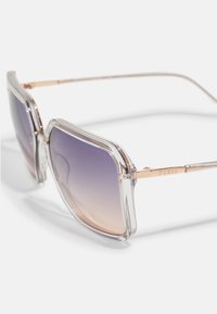 Lunettes de soleil surdimensionnées transparentes avec des verres dégradés violet, une monture claire et des accents métalliques dorés. Logo gravé sur la monture.