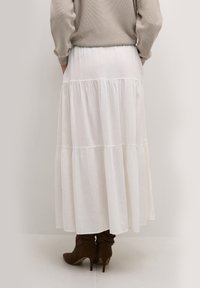 Bílá vrstvená maxi sukně vyrobená z lehkého materiálu, s volným designem a jemnými švy, v kombinaci s hnědými vysokými botami.