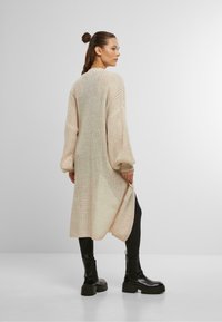 Strickjacke in Creme mit lockerer Passform, langen Ärmeln, offener Vorderseite und längerem Rückenbund, kombiniert mit schwarzen Leggings und Stiefeln.
