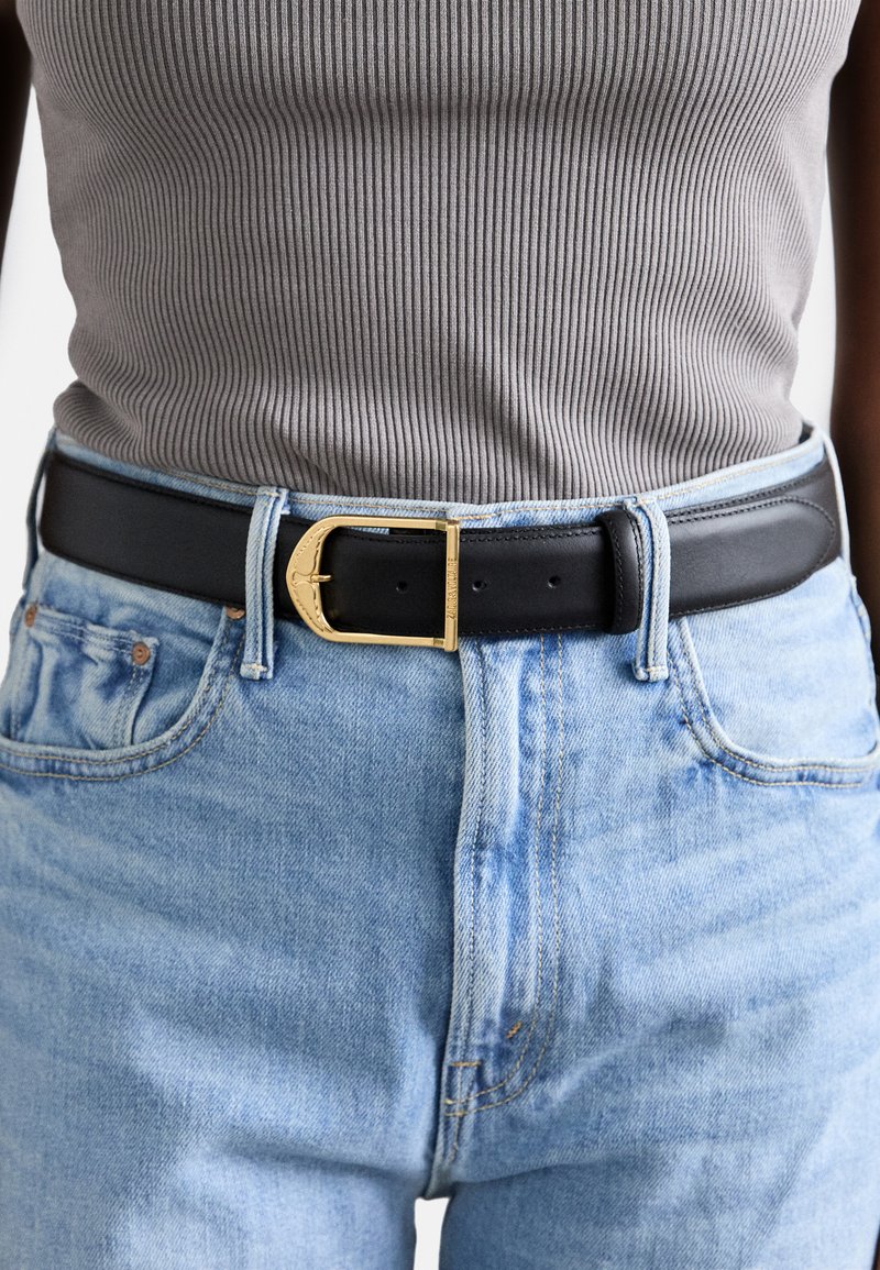 Zwarte leren riem met een gouden gesp, gedragen over lichtblauwe denim jeans, vergezeld van een grijze geribde tanktop.