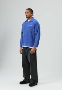 Blauwe, pluizige trui met een kraag en lange mouwen, uitgevoerd in een gestructureerde stof met witte geborduurde letters, gecombineerd met losse zwarte jeans.