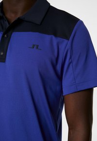 Blaues Poloshirt mit schwarzem Kragen und Schulterpaneelen. Glatte Textur, kurze Ärmel, drei Knöpfe und ein kleines schwarzes Logo auf der Brust.