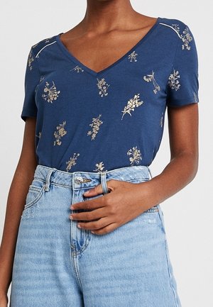Vrouw draagt een blauw T-shirt met korte mouwen en V-hals met kleine bloemenprints en lichtblauwe jeans met hoge taille en knoopsluiting.