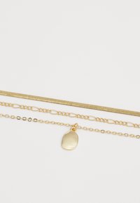 Collier à plusieurs couches en ton doré comprenant trois chaînes : une chaîne serpent, une chaîne à maillons longs et une chaîne avec un pendentif ovale.