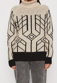 Stickad tröja med hög polokrage, beige botten och svarta geometriska mönster. Har ribbade ärmslut och nederkant, samt en avslappnad passform.