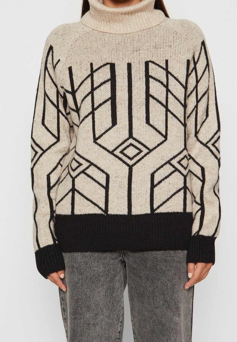 Stickad tröja med hög polokrage, beige botten och svarta geometriska mönster. Har ribbade ärmslut och nederkant, samt en avslappnad passform.