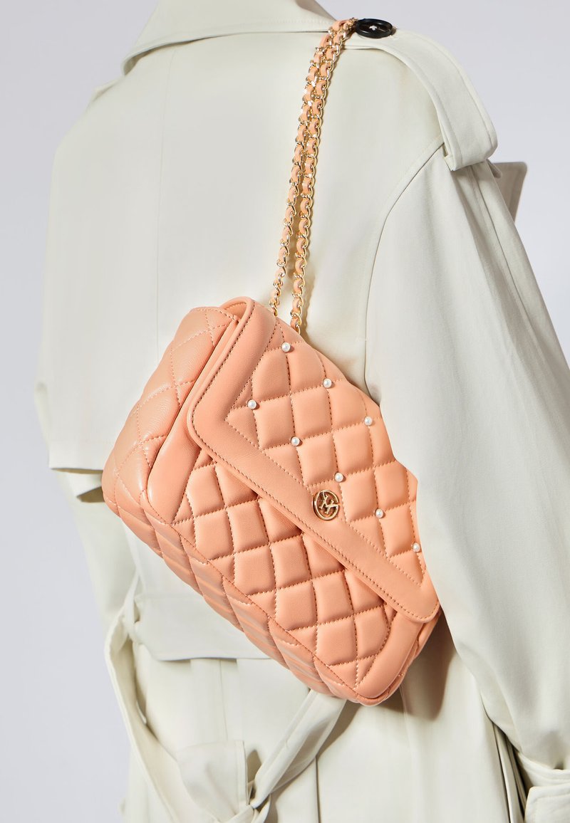 Brzoskwiniowa torebka pikowana z łańcuszkiem, ozdobiona małymi białymi perełkami i detalem z logo, zaprezentowana na tle jasnego płaszcza.