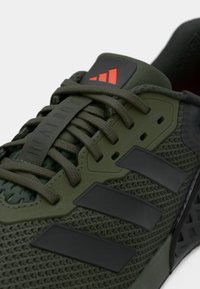 Baskets de sport vert olive avec un dessus en maille, accents noirs, et trois rayures noires. Elles possèdent des lacets plats olive et un système de laçage ajustable.