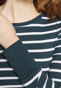 Randig långärmad skjorta med en teal-färgad bas och vita horisontella ränder. Har rund halsringning och åtsittande ärmslut. Mjuk, stretchig tyg.