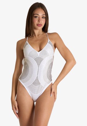 Giovane donna con lunghi capelli castani che indossa un costume intero bianco con motivi di ritagli circolari e sottili spalline.