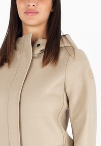 Giacca beige con cappuccio realizzata in materiale morbido con una texture liscia, caratterizzata da un accento logo discreto sulla manica e una chiusura con zip.