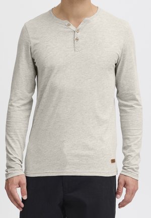 Chemise henley manches longues gris clair avec trois boutons au col portée par une personne portant un pantalon noir, les mains détendues le long du corps.
