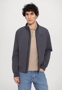 Grå zip-jakke med ståkrage, svart trim og EA7-logo. Kombinert med en beige polo-skjorte og blå jeans. Glatt tekstur.
