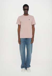 Helles rosa Baumwoll-T-Shirt mit kurzen Ärmeln und kleinem Logo, kombiniert mit ausgewaschenen blauen Jeans und weißen Turnschuhen.