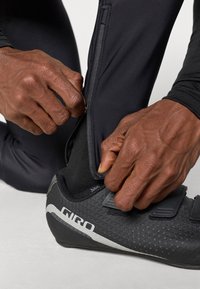 Čierne cyklistické nohavice so zipsom na členku, nosené cez čierne cyklistické topánky s textúrovaným povrchom a suchými zipsami.