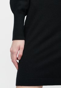 Ruka odložená vedľa čiernych dlhých rukávov pletených šiat, ukazujúca textúru látky a detaily manžiet na jednoduchom pozadí.