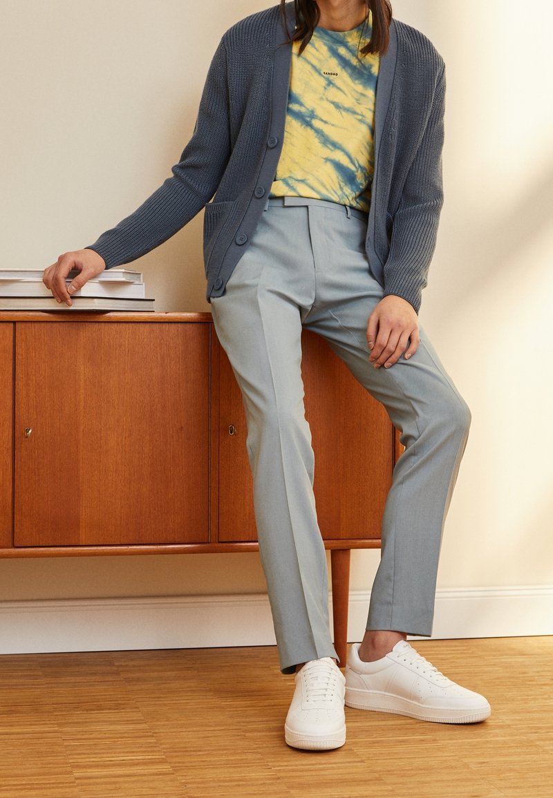 Ljusblå stickad cardigan över en gul och blå tie-dye t-shirt, kombinerad med ljusgråbyxor och vita sneakers, mot en träbakgrund.