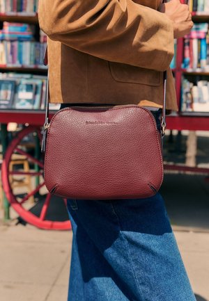 Person iført blå jeans og brun jakke bærer en tekstureret vinrød crossbody taske med lynlås på et udendørs marked.
