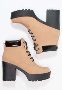 Botas de tobillo de ante sintético beige con un tacón de plataforma negra, que presentan cordones negros y un acento negro brillante en el cuello.