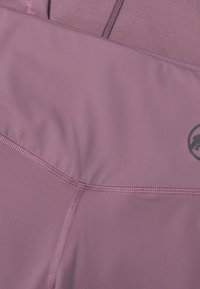 Lys lilla atletisk stof med en glat tekstur, der har kontrasterende syninger og et cirkulært logo i nederste højre hjørne.