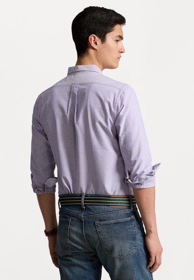 Ανοιχτό μοβ πουκάμισο με μακριά μανίκια, γυρισμένα μανίκια και πιέτα στο πίσω μέρος, φορεμένο με μπλε τζιν και ριγέ ζώνη. Εμφανίζεται η πίσω όψη.