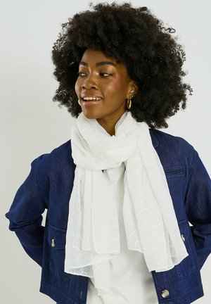Mujer con cabello rizado que lleva pendientes de aro dorados, chaqueta azul oscuro y pañuelo blanco con estampado, mirando hacia un lado con una leve sonrisa.