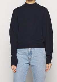 Maglione lavorato a maglia blu navy con collo alto, spalle scese e maniche lunghe, abbinato a jeans azzurri. Tessuto morbido e vestibilità rilassata.