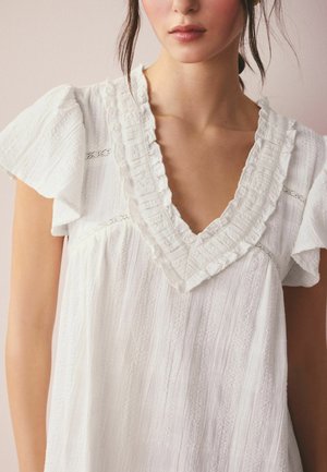 Moteris vilki baltą tekstūruotą suknelę su giliais V formos iškirpte ir raukiniais rankovėmis prieš vientisą foną.