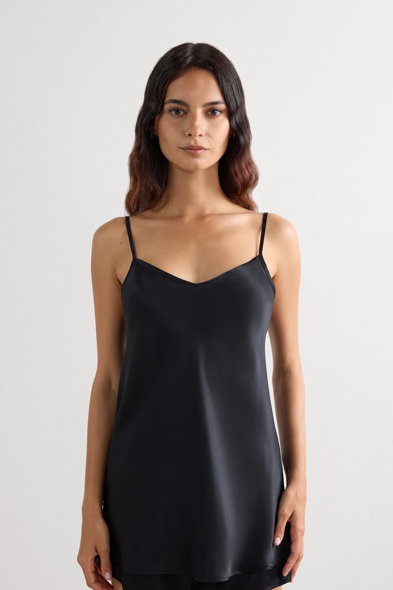 Femme aux longs cheveux ondulés portant une robe nuisette noire en satin à fines bretelles, debout devant un fond uni gris clair.