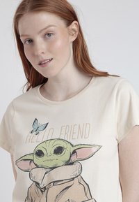 Camiseta color beige claro con un personaje de dibujos animados que tiene grandes ojos, una cara verde y una capa marrón, acompañada del texto "HOLA AMIGO" y un gráfico de mariposa.