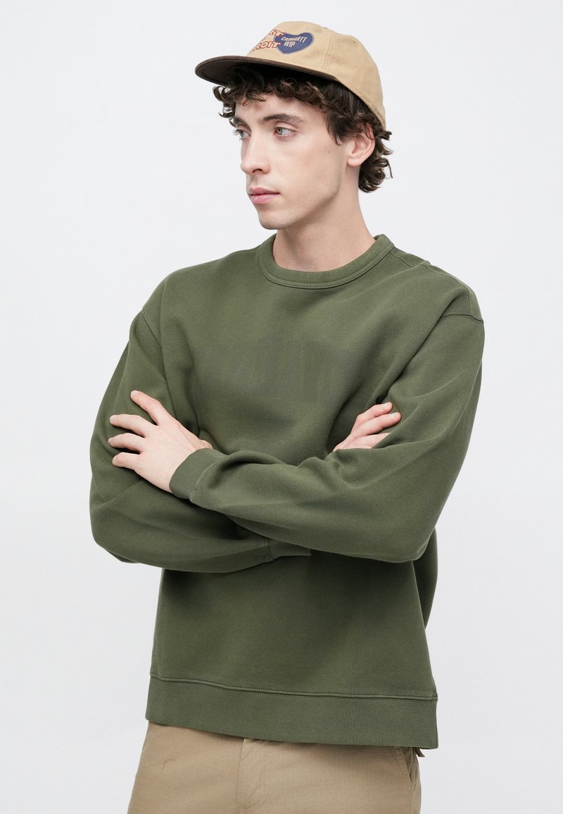 Olive green sweatshirt with a crew neckline and ribbed cuffs, featuring a subtle logo print. A beige cap with a graphic logo completes the look.