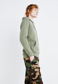 Khaki zielona bluza z kapturem z kieszenią z przodu, połączona z spodniami cargo w kamuflażu z dużymi bocznymi kieszeniami i teksturowanym materiałem.