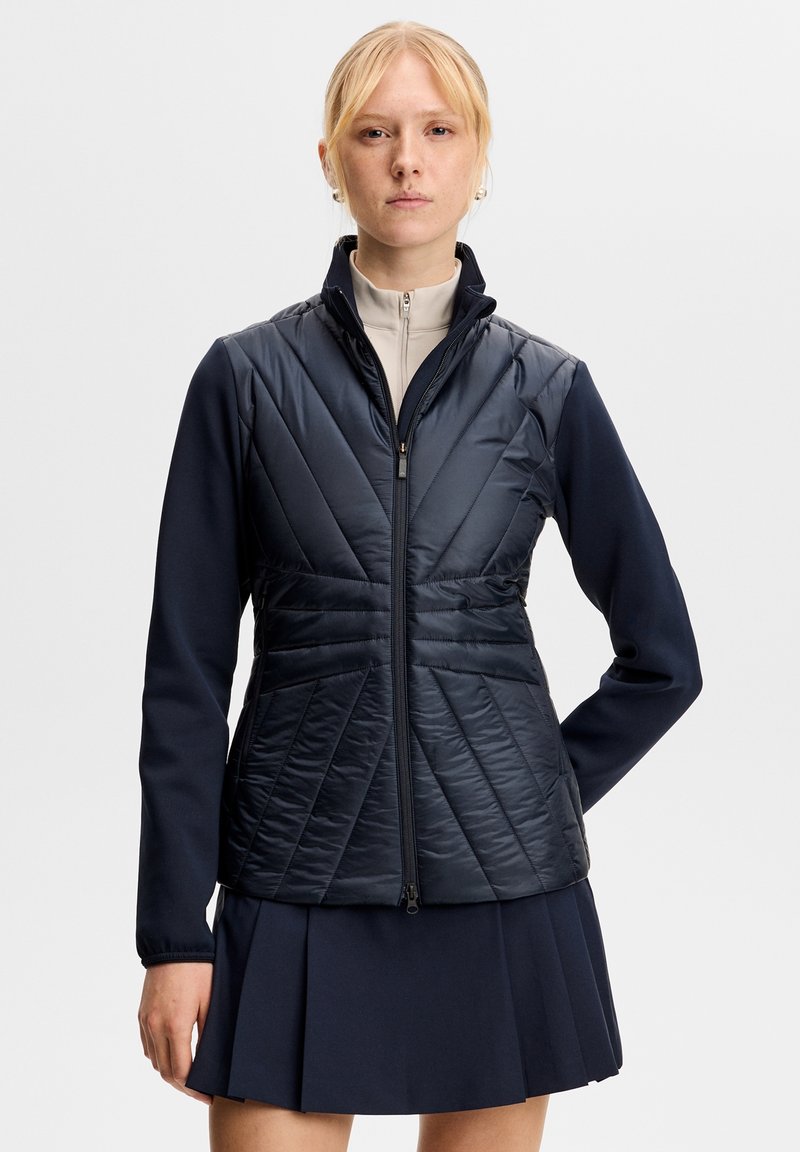 Blond kvinde står fremadrettet iført en marineblå quiltet jakke med lynlås over en beige top og en navy plisseret nederdel mod en ensfarvet baggrund.