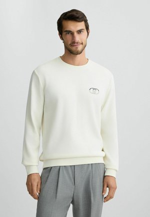 White sweatshirt made of soft fabric with a round neckline, ribbed cuffs, and a small logo on the left chest, paired with gray pleated trousers.