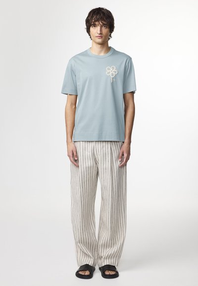Lys blå bomulds t-shirt med broderet blomsterdesign, parret med cremefarvede stribede bukser. Model iført sorte sandaler.