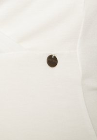 Tecido branco com uma textura suave, apresentando uma pequena etiqueta redonda em bronze que diz "LOVE+WANT" anexada à costura.