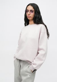 Νεαρός/νέα με μακριά σκούρα μαλλιά, φορώντας ανοιχτό μωβ φούτερ, γκρι φόρμες και διαφανή μωβ γυαλιά ηλίου, με τα χέρια στις τσέπες, ουδέτερη φόντο.