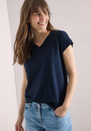 Femme souriante aux longs cheveux bruns portant un t-shirt bleu marine à manches courtes et col en V, rentré dans un jean bleu clair, une main dans la poche.