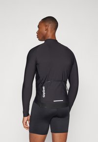 Black long-sleeve cycling top, fitted design, smooth texture, features a rear pocket and reflective accents. Worn with black shorts.