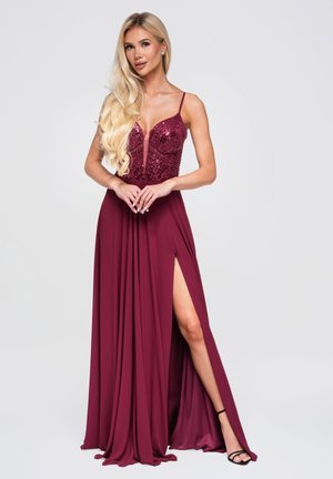 Femme aux longs cheveux blonds portant une robe de soirée bordeaux sans manches avec un corsage à sequins et une fente haute sur la cuisse, sandales noires, sur fond clair.