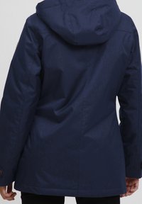 Marinblå, lätt regnjacka med huva, texturerad tyg och knappdekorationer på ärmarna. Har en rak skärning och sömdetaljer på baksidan.