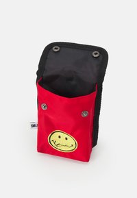 Röd tygpåse med svart foder, med en gul smiley-ansiktsdesign och tryckknappar framtill. Texturerad utsida.